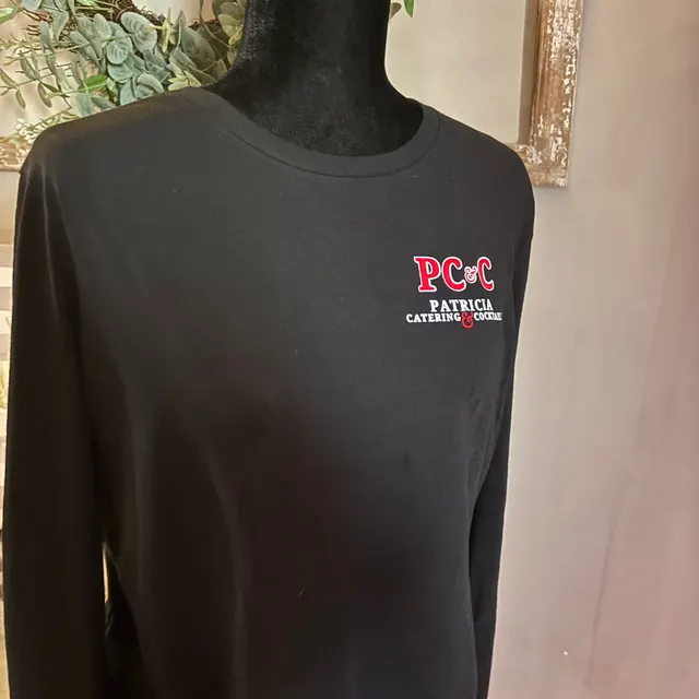 Black long-sleeve shirt on a mannequin torso reading PC&C, PATRICIA, CATERING & COCKTAILS, with an ornate gold frame on the wall behind