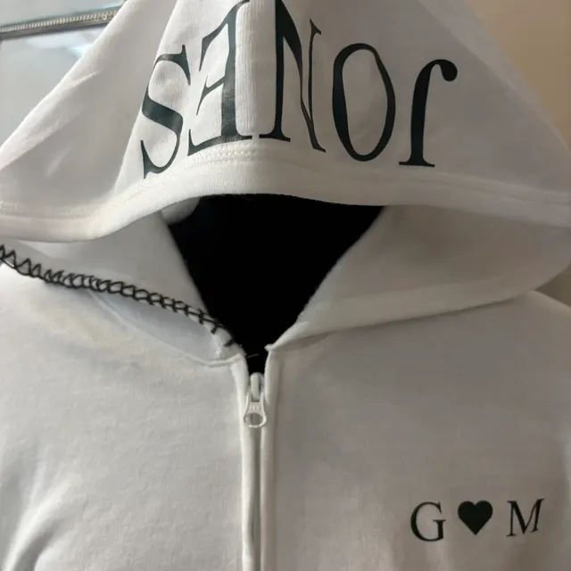 White full-zip hoodie with a black-lined hood reading SENIOR and G-heart-M lettering on the chest, the heart standing in for the A in GAM