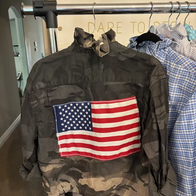 Camo jacket with an American flag patch, hanging on a rack beside plaid shirts