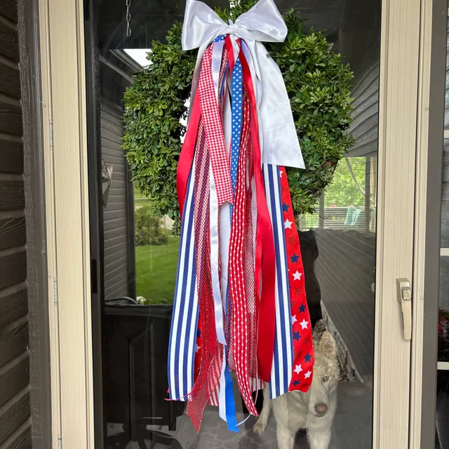 Boxwood wreath with a large white bow and cascading red, white and blue star-and-stripe ribbons, hanging on a glass door