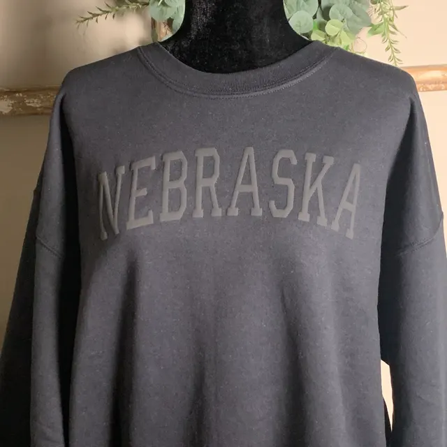 Grey sweatshirt on a black mannequin torso with NEBRASKA arched in light grey varsity letters, greenery in the background