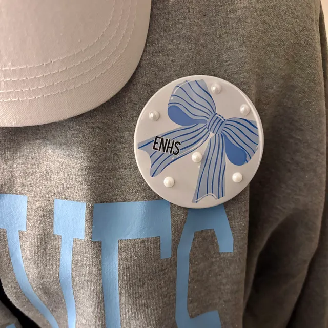 Elkhorn North spirit set laid flat — grey shirt with light blue lettering, a white cap, and a circular ENHS pin with a blue striped bow