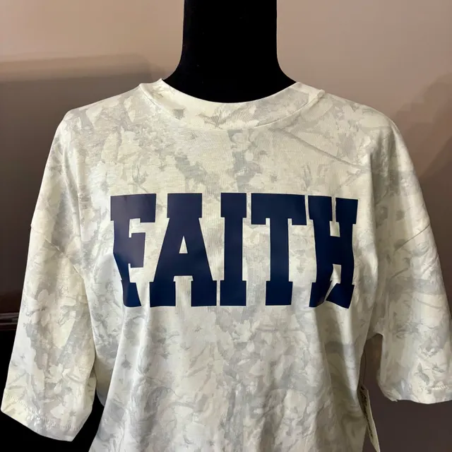 White and grey abstract-pattern t-shirt on a black mannequin with large dark blue block letters spelling FAITH across the chest